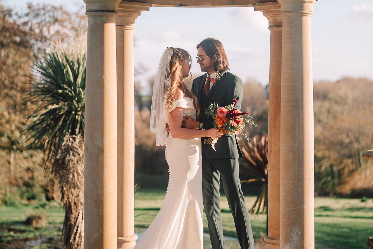 Tredudwell Manor autumnal wedding bouquet designed by Trelowen Flowers. Couple in wedding attire standing under a stone archway with a garden background
